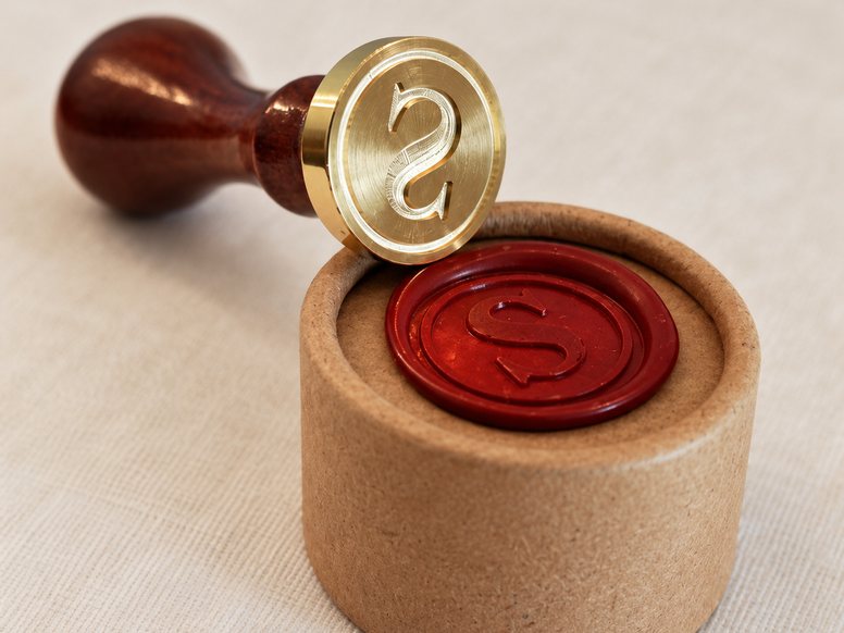 Brass seal die with a turned wooden handle, its mirror-cut S facing the camera, beside a wax impression.